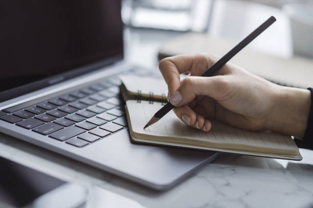 istock Tight focus on a woman's hand as she writes in a notepad on a contemporary laptop, background subtly blurred 1499801767