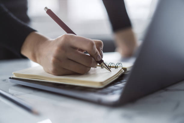 The image captures a woman's hand in close view as it writes in a notepad on a chic laptop, background out of focus