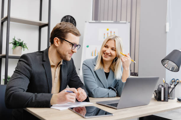 istock Cheerful business people using a laptop in an office. Young entrepreneurs smiling while working together in a modern workspace. Two young business people sitting together at a table. 1499803746