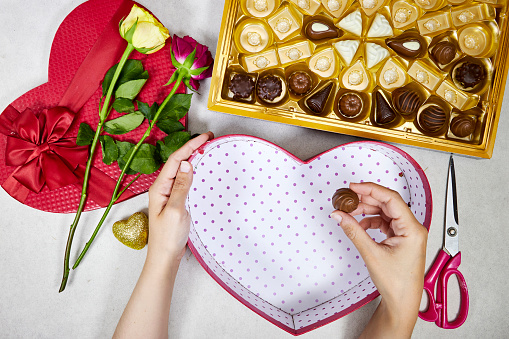 chocolate pieces from various type in box for valentine's day with woman putting pieces in open red heart shaped box and roses flowers