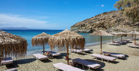 Straw umbrella, lounger in row on sandy empty beach summer sunny day. Relaxation at seaside chair under parasol, Tinos island Cyclades Greece.
