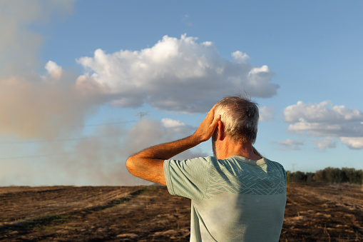 Close-up of man who is on his back and has his hands on his head looking at his burned field after fire. There is smoke in background of image.