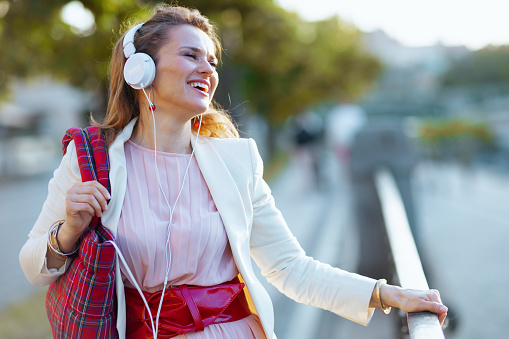 happy elegant middle aged woman in pink dress and white jacket in the city with headphones and red bag.