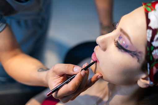 Makeup artist is painting contour lips of a young woman with a pencil.