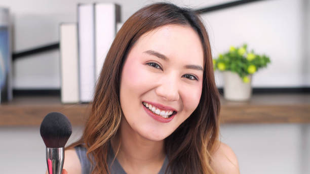 행복한 기뻐하는 예쁜 아시아 여성의 초상화는 얼굴에 붓 홍당무를 들고 카메라에 미소를 짓고 있다. 메이크업 방법을 보여주는 아름다운 여성 블로거. 뷰티 블로거, 온라인 인플루언서 컨셉 - statue human face women human skin 뉴스 사진 이미지