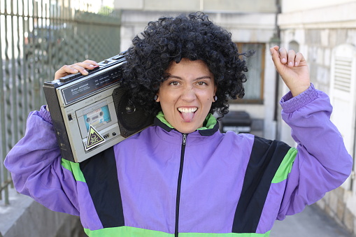 A gorgeous Hispanic woman in her twenties is listening to music on the street. She uses a vintage stereo cassette and wears a colorful outfit.