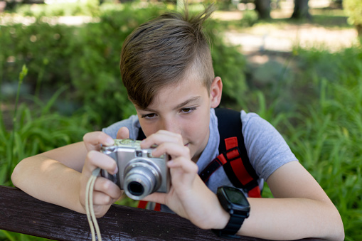 Young photographer with backpack. Boy holding a digital camera and taking photos in the park.