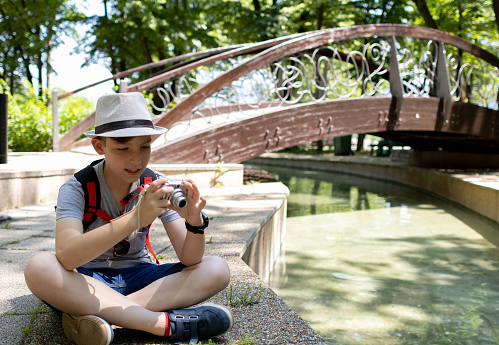 Young photographer with backpack and hat. Boy holding a digital camera and taking photos in the park.
