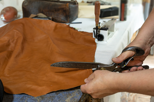 Craftsman cutting leather with big old scissors in his workshop, material to create bags, belts, clothing or upholstery copy space, selected focus, narrow depth of field