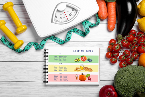 Information about grouping of products under their glycemic index. Notebook, measuring tape, fruits, vegetables, dumbbells and floor scale on white wooden table, flat lay