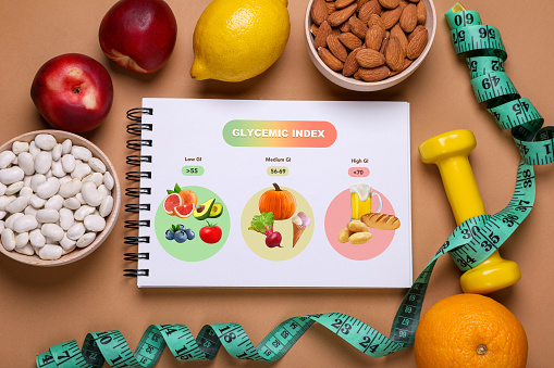 Information about grouping of products under their glycemic index. Notebook, measuring tape, dumbbell, beans, almonds and fruits on light brown background, flat lay