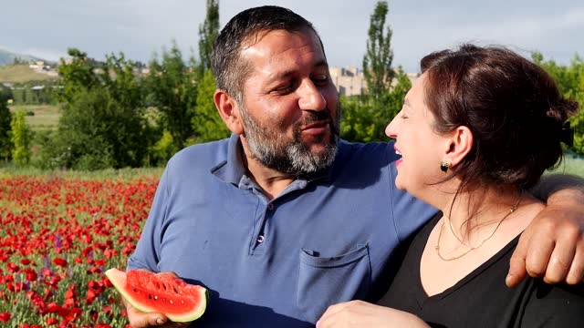 Very cute and happy adult middle eastern husband and wife biting watermelon together at the same time holding a piece of juicy fruit in their hands outdoors in a field with red flowers at sunset