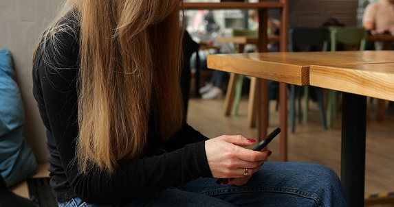 Unrecognizable young woman in coffee shop waiting for order with phone on knees. Faceless female with long brown hair sits at empty table type on smartphone, cars and people in street in background.
