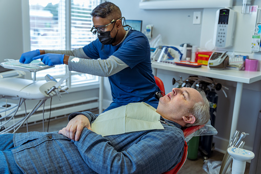 A caucasian man lies in a dental chair with a concerned expression on his face while in the background his male dentist of Indian descent organizes sterile instruments in preparation of performing a dental procedure.