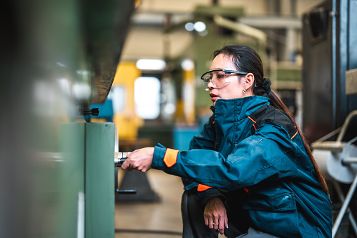 Mid adult female blue collar worker operating an industrial machine in a factory. She is wearing protective clothes and eyeglasses. Adjusting machine settings.