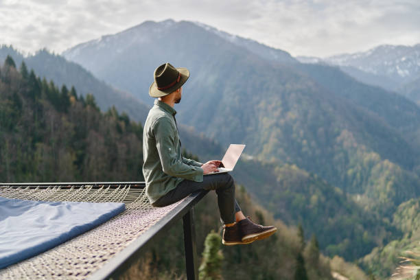 istock Young man freelancer traveler wearing hat anywhere working online using laptop and enjoying mountains view 1499876087