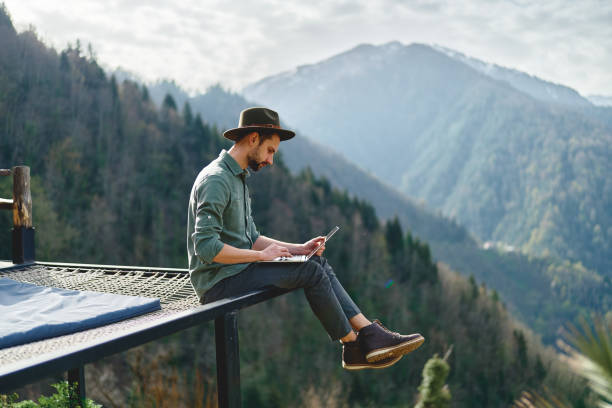 Young man freelancer traveler wearing hat anywhere working online using laptop and enjoying mountains view