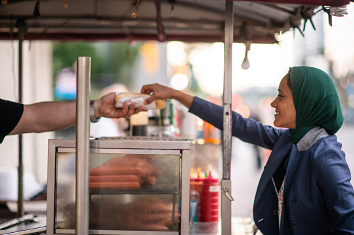 A young Muslim woman purchases a hot dog from a street vendor as she ventures outside her office building for her lunch break.  She is dressed professionally in a suit and Hijab.
