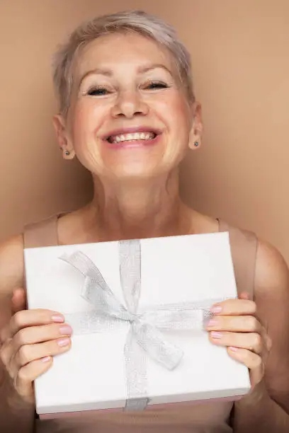 Photo of smiling lady pensioner with short hair holding present box isolated beige color background