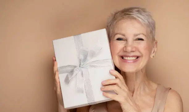 Photo of smiling lady pensioner with short hair holding present box isolated beige color background