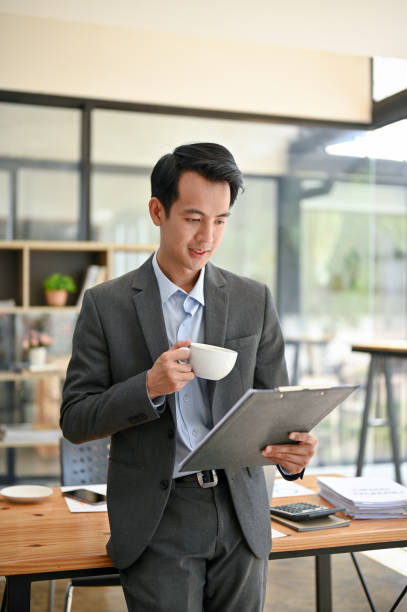 istock A smart Asian businessman reading document and sipping coffee while standing in the office. 1499893952