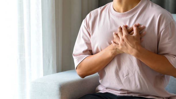 Young man sitting on sofa in the living room using both hands holding chest with symptom heart attack disease.He was worried because of the pain on the left side of his chest. stock photo
