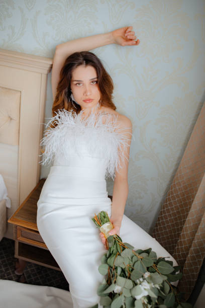 istock Young lady in wedding dress with feathers leaning on wall while sit on bedside table. Bride holding bouquet of flowers. 1499913225