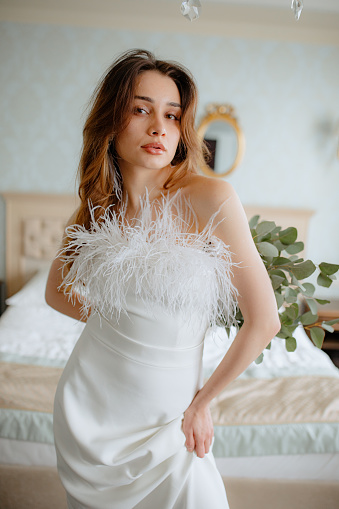 Delightful young brunette in wedding dress with little and fluffy feathers standing in front of bed in hotel room. Confident lady holding bouquet of flowers behind back with hand.