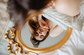 Brunette lying on bed and looking at gold-rimmed mirror. Bride holding pillow under hands and admiring reflection.
