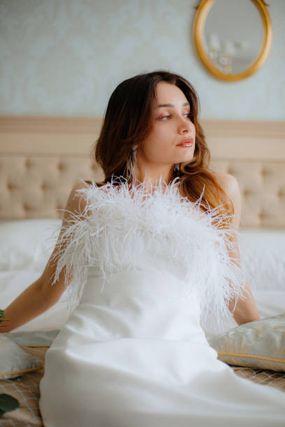 istock Confident woman in wedding gown decorated with feathers leaned back on hands while sitting on bed. Bride looking aside. 1499915902