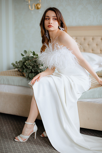 Loveable young woman with brown hair sitting on bed in shoulderless wedding dress adorned with feathers and high heels shoes. Bride with tassel earrings looking aside, bouquet of flowers lying near.
