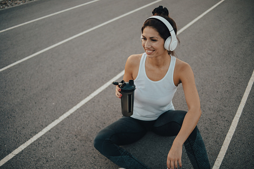 Sportswoman rests after training, sits on the track in the stadium and listens to music on wireless headphones, training props are next to her