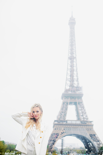 Charming lady with flowing blond hair and in white clothes softly touching face with hand against backdrop of famous Eiffel Tower. Adult girl walking in Paris next to popular place.