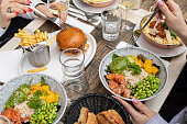 Girls with forks in hands trying food in cafe. Salads of fish, rice and veggies, burger, french fries lying on table.