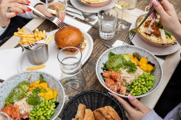 meninas com garfos nas mãos experimentando comida no café. saladas de peixe, arroz e legumes, hambúrguer, batatas fritas deitadas na mesa. - foodie - fotografias e filmes do acervo