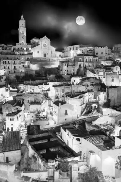 A suggestive night scene with the moon of the historic heart of Matera, in southern Italy, known worldwide as the 'Sassi' (Matera's Stones). Top left, the splendid facade of the Cathedral in Apulian Romanesque style. The ancient city of Matera, in the region of Basilicata, in southern Italy, is one of the oldest urban settlements in the world, with a human presence that dates back to more than 9,000 years ago, in the Paleolithic period. The Matera settlement stands on two rocky limestone hills called 'Sassi' (Sasso Caveoso and Sasso Barisano), where the first human communities lived in the caves of the area. The rock cavities have served over the centuries as a primitive dwelling, foundations and material for the construction of houses, roads and beautiful churches, making Matera a unique city in the world. In 1993 the Sassi of Matera were declared a World Heritage Site by Unesco. Image in high definition quality.