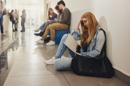 Sad high school female student sitting on a floor alone during a break in a hallway. Copy space.