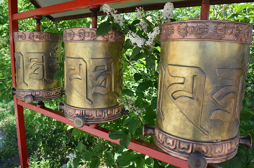 Three brass and copper Buddhist prayer wheels surrounded by greenery and lilac bushes
