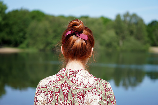 back view of unrecognizable young woman with floral dress and red hair bun looking at lake in solitude