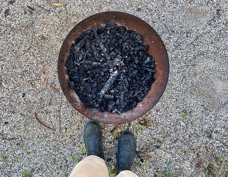 Horizontal flat lay looking down to outdoor rusted round fire pit with charcoal burnt timber log embers with black boots and tan pants on gravel ground at country rural property Australia