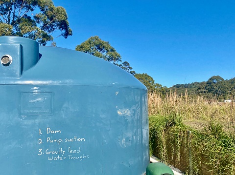 Horizontal close up of large blue plastic country farm water tank against Australian bush landscape with white writing on tank in rural area