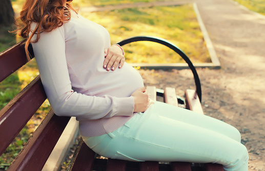 Happy young pregnant woman sitting on bench in summer park