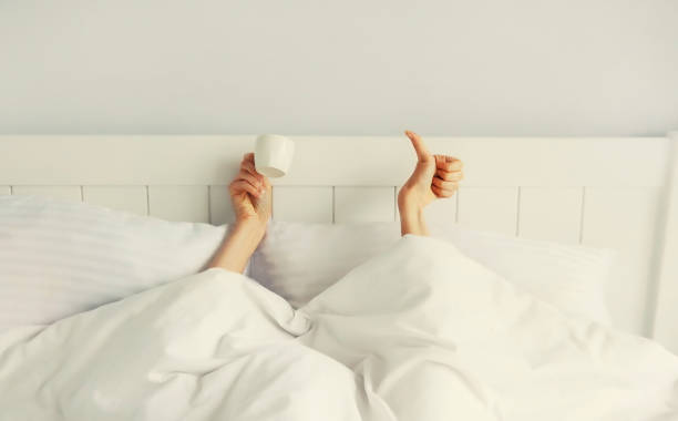 istock Cheerful lazy woman waking up after sleeping lying in soft comfortable bed showing empty cup coffee stretching her hands up from under the blanket in white bedroom 1499939002