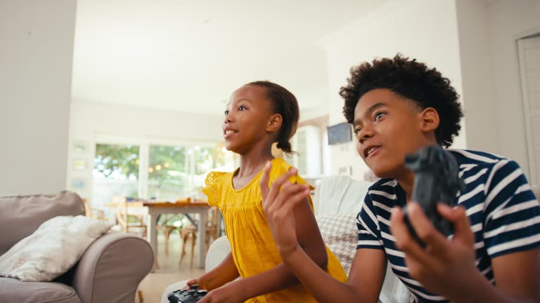 Brother And Sister Sitting On Sofa At Home Playing Video Game Together With Girl Winning