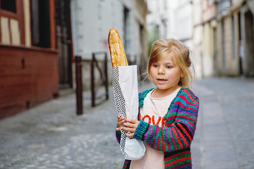 Adorable little preschool girl with fresh French baguette on the street side of the city. Happy small child in Paris, France