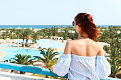 A young attractive girl looks down on the hotel complex on the first coastal line of the sea. Beautiful green area with pool and places for relaxation. The concept of tourism, vacations
