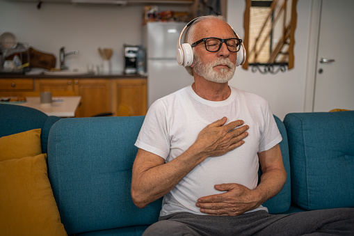 Mature senior man sitting on bed practicing guided meditation at home, hands resting on chest and stomach, squinting and relaxing