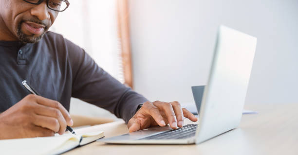 istock Closeup businessman hand holding pen writing on notebook, work on laptop computer in modern office. Student lecturing while studying online internet course. Write on journal diary banner (blur) 1499945710