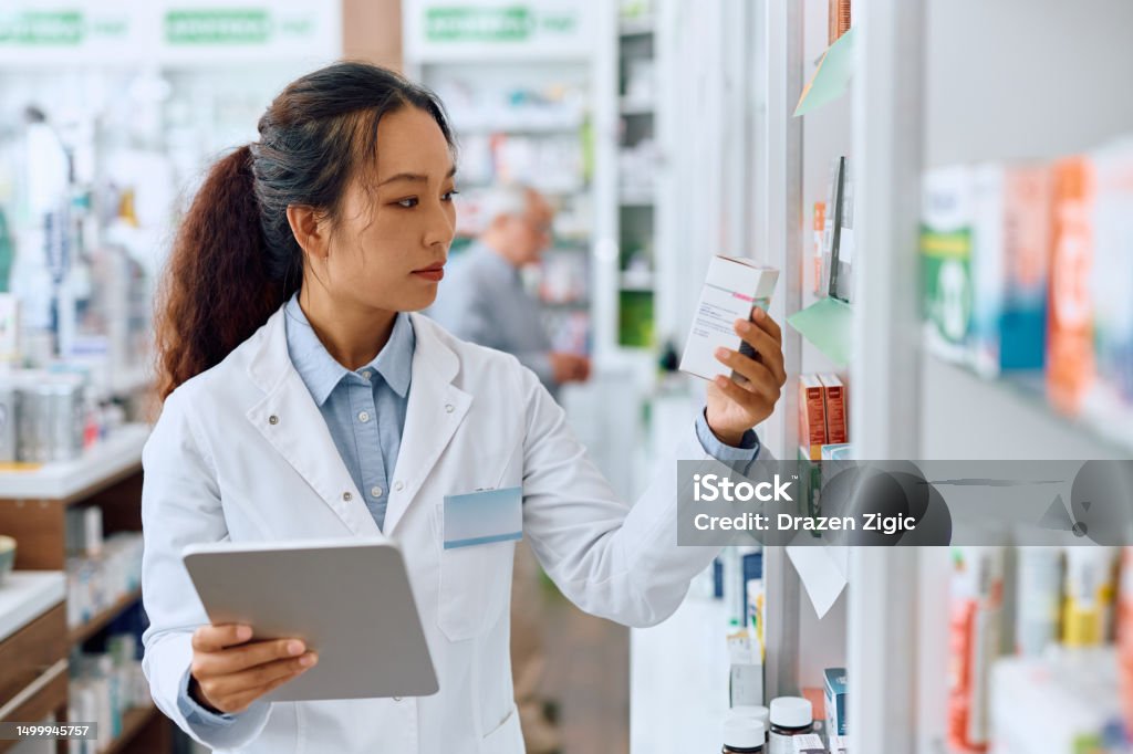 Female Chinese pharmacist using digital tablet while working in a pharmacy. Young Chinese pharmacist using touchpad while checking a medicine inventory in drugstore. Pharmacist Stock Photo