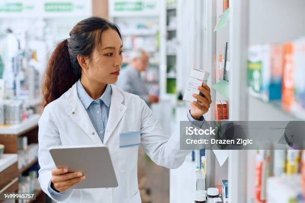 Female Chinese Pharmacist Using Digital Tablet While Working In A Pharmacy Stock Photo - Download Image Now